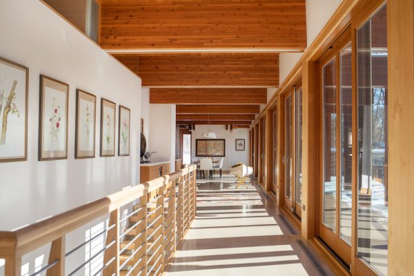 A wall of windows along the front of the home flood the main floor with winter sunlight, highlighting the wooden beams on the ceiling as well as the homeowner’s collection of art.